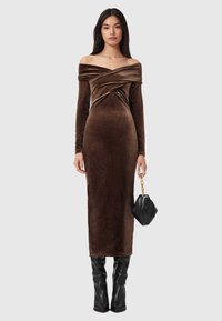 Brązowa aksamitna sukienka odsłaniająca ramiona, z długimi rękawami, marszczeniem na biuście i dopasowaną sylwetką. Zestawiona z czarną torebką.