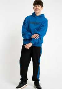 Nastolatek z krótkimi kręconymi włosami, uśmiechający się, ubrany w niebieską bluzę z napisem, czarne spodnie z niebieskimi paskami i czarne sneakersy, stojący na białym tle.