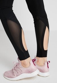 Leggings noirs avec des découpes en maille transparente au niveau des mollets, offrant un ajustement moulant et des coutures contrastantes. Associés à des chaussures de sport rose clair.