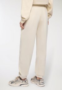 Pantaloni da tuta color crema con una vita elastica, polsini affusolati e una texture morbida, abbinati a sneaker beige e grigie.