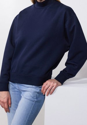 Persoon leunend op een wit oppervlak, gekleed in een marineblauwe top met lange mouwen en colletje en lichtblauwe spijkerbroek, tegen een effen witte achtergrond.