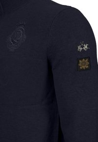 Navy blue knitted pullover with a high collar. Features embossed logo, two embroidered patches on the sleeve, and a textured body.