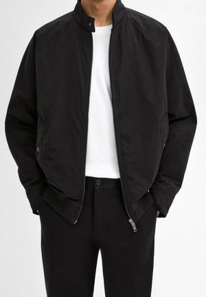 Homme portant une veste noire à fermeture éclair sur une chemise blanche et un pantalon noir, debout les mains dans les poches de la veste.