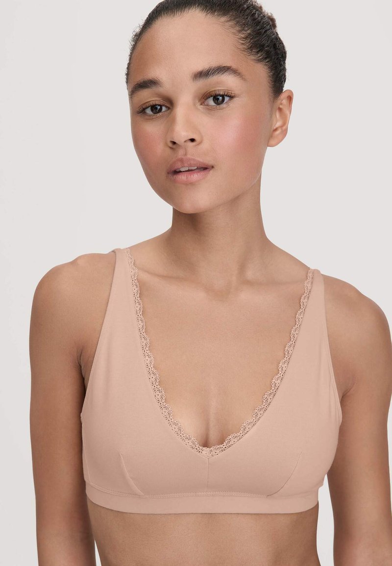 Jonge vrouw met krullend haar dat naar achteren is vastgebonden, draagt een huidkleurige, kantafgewerkte zachte beha tegen een eentonige achtergrond.