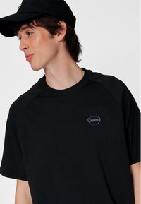 Svart T-shirt med korta raglanärmar och en liten blå logotyp. Tyget ser mjukt och lätt ut. Modellen bär en svart keps.