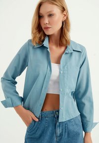 Lichtblauw overhemd met lange mouwen, met knopen, een kraag en een borstzakje. Zacht stof, iets losvallend model, gecombineerd met high-waisted jeans.