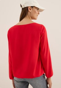 Röd långärmad blus i lätt material med löst sittande passform, med en vid halsringning och mjukt veckade ärmar, tillsammans med ljusgrå jeans.