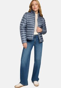 Veste matelassée bleu clair et légère avec protège-menton, épaules structurées et fermeture éclair. Associée à un jean à jambes larges et des chaussures blanches.