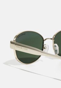 Sluneční brýle ve zlatém odstínu s kulatými rámy, zelenými skly a vyrytým textem "MICHAEL KORS" na nožkách. Obsahují texturované akcenty na ramenou.