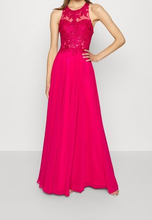 Kvinde iført en ærmeløs, knaldrød gulvlængde kjole med blomsterblonder bodice og flydende chiffon skørt, stående med den ene hånd på hoften.