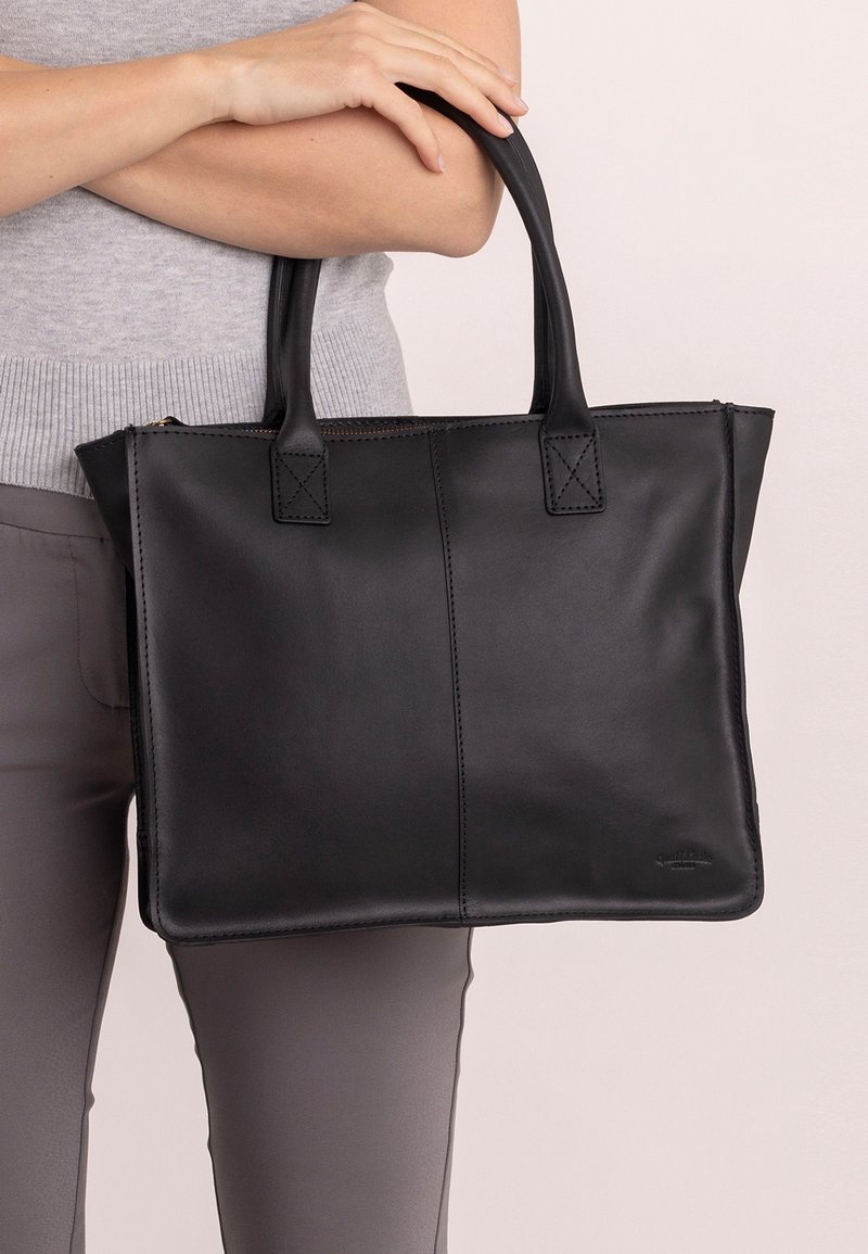 Personne tenant un grand sac cabas en cuir noir avec double poignée, portant un haut sans manches gris et un pantalon taupe.
