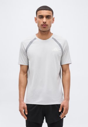 Junger Mann mit einem hellgrauen kurzärmeligen Sportshirt und schwarzen Shorts, stehend mit entspannten Armen vor einem einfarbigen Hintergrund.