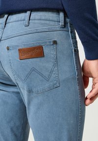Jeans de mezclilla azul claro con un parche de cuero marrón de Wrangler, que cuentan con bolsillos traseros con un patrón de zigzag y remaches plateados.