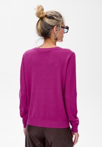 Kobieta z blond włosami upiętymi w kok nosi okulary w muszelkowym wzorze, kolczyki w kształcie kół, jasnoróżowy sweter i ciemne spodnie, stojąc tyłem na białym tle.