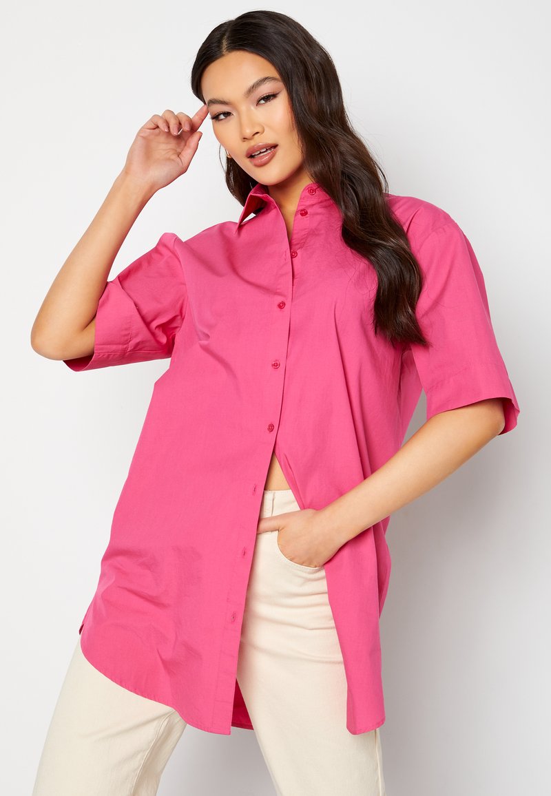 Camicetta rosa a maniche corte con colletto a punta e vestibilità dritta, con spacchi laterali e texture di cotone morbido. Indossata con pantaloni beige chiaro.