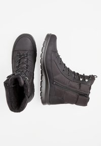 Botas de tobillo negras, aisladas, con paneles texturizados, diseño de cordones, cremallera lateral y suela de goma. Forro suave visible en la parte superior.