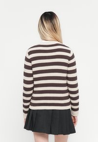 Person med langt blondt hår iført en brun og hvit stripete genser og et svart plisserte skjørt, vendt bort fra kameraet mot en hvit bakgrunn.