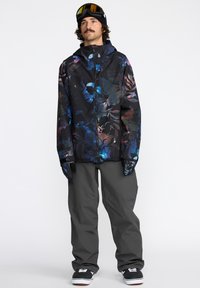 Veste florale noire pour hommes avec des motifs bleus, violets et roses, associée à un pantalon gris et des gants. Les accessoires comprennent des lunettes de neige.