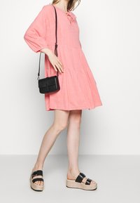 Rosa Leinenkleid mit dreiviertel Ärmeln, einem Bindedetail am Ausschnitt und einem gestuften Design. Schwarze Umhängetasche und schwarze Plateau-Sandalen.