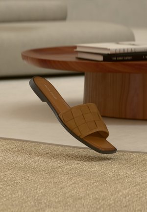 Sandale mule en cuir tressé marron reposant à côté d'une table basse ronde en bois avec des livres empilés sur un tapis beige.