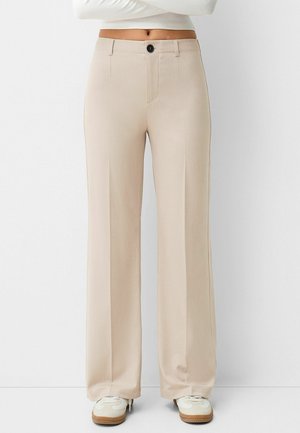 Personne portant un pantalon large taille haute beige avec un bouton noir et des baskets blanches, debout devant un fond blanc uni.