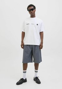 Hvid oversized t-shirt med sorte grafiske prints, grå plisserede shorts, sorte sko og hvide sokker med sorte striber.