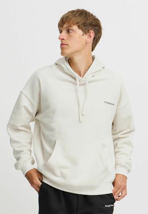 Cream hoodie with a front pocket, drawstrings, and logo on the chest. Fabric has a smooth, soft texture. Paired with black pants.