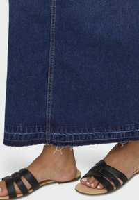 Mörk jeanskjol med fransig nederkant och kontrasterande sömmar, kombinerad med svarta platta sandaler med knutdetalj.