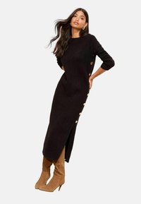 Rochie neagră tricotată cu mâneci până la trei sferturi, accente cu butoni pe lateral și o fanta laterală. Asortată cu cizme din suede de culoare bej, până la genunchi.
