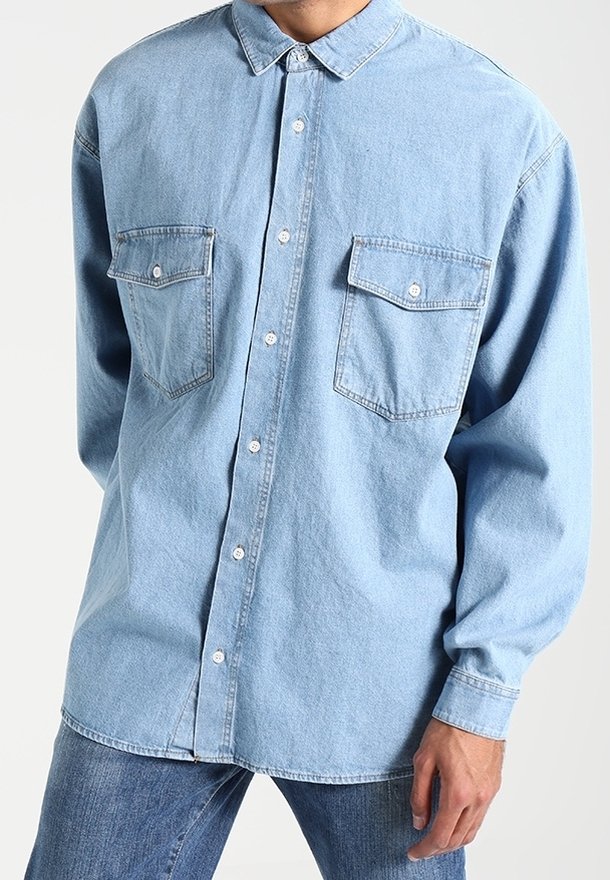 Jasnoniebieska koszula dżinsowa z zapięciem na guziki i kieszeniami na klapie, noszona na wierzchu z niebieskimi dżinsami, ukazująca górną część torsu i prawą rękę.