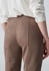 Braune, samtähnliche, figurbetonte Hosen mit Rückennaht-Detail, kombiniert mit einer weißen Bluse; das Bild konzentriert sich auf den unteren Rücken und die Passform der Hose.