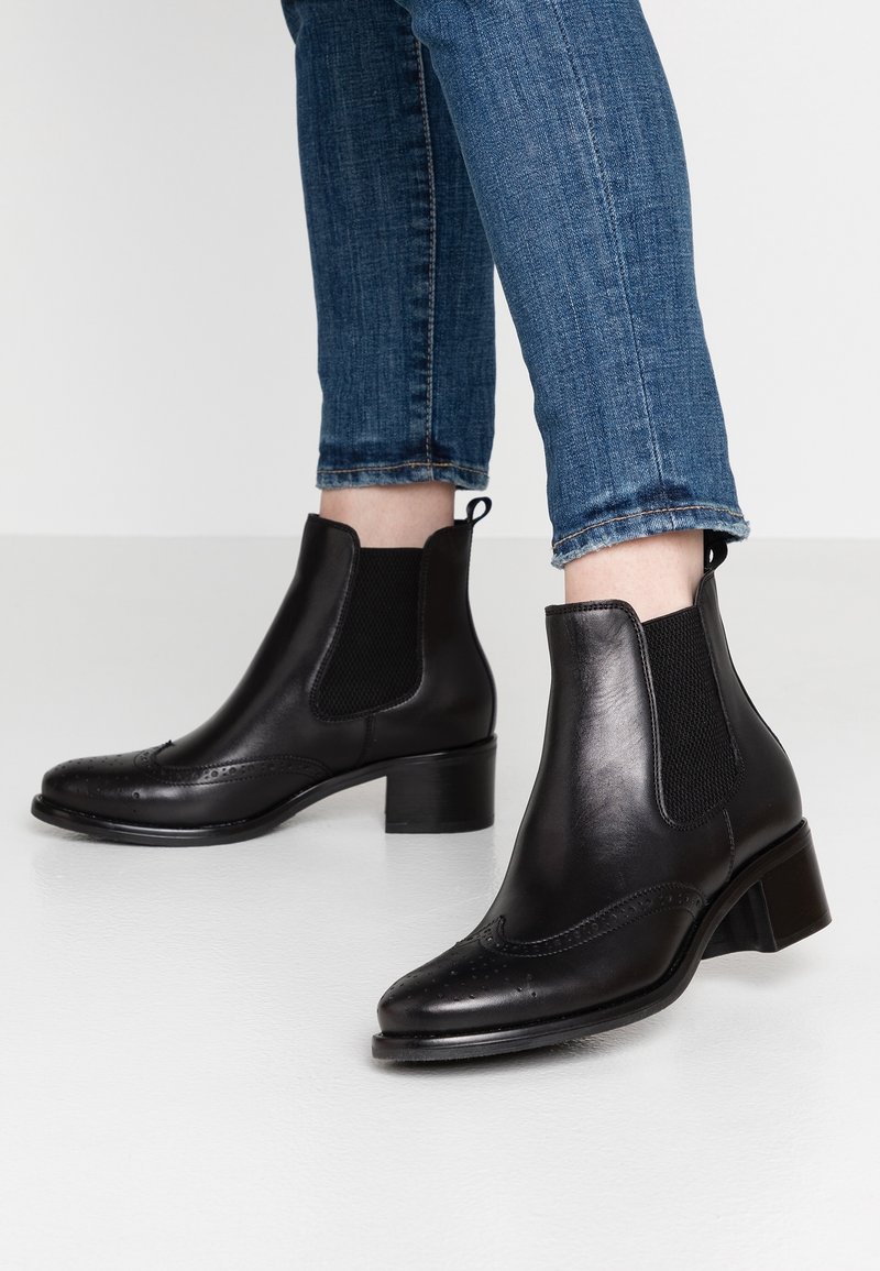 Bottines chelsea en cuir noir avec des panneaux élastiques sur les côtés, un bout pointu et des détails décoratifs en brogue. Talon épais et finition texturée.