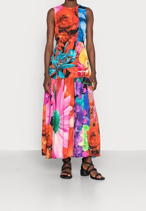 Kobieta w bezrękawnikowej, wielokolorowej sukience maxi z kwiatowym wzorem patchworkowym, nosząca czarne sandały na słupku z paskami, stojąca na białym tle.