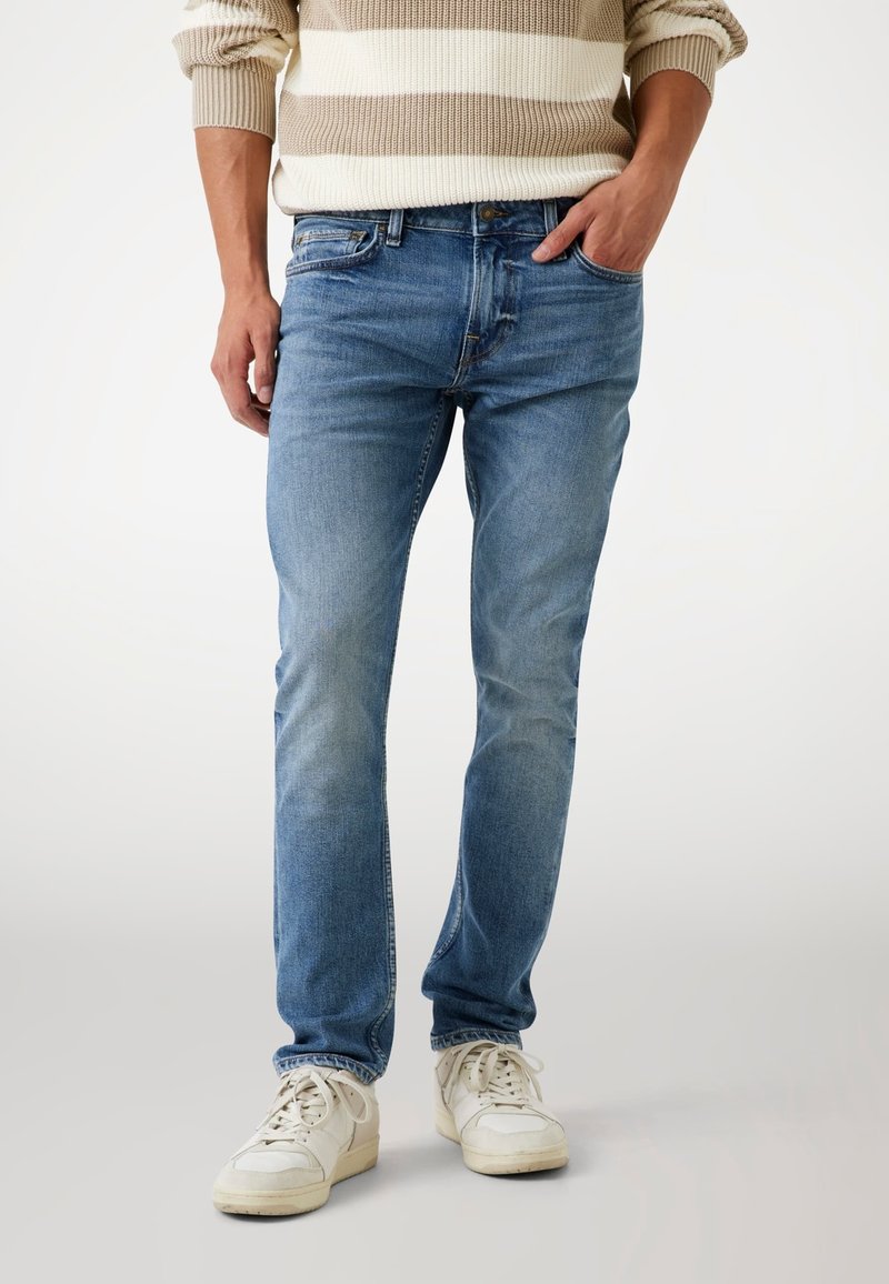 Jean en denim light blue slim fit avec texture délavée, design classique à cinq poches et fermeture à bouton, associé à des baskets blanches.
