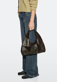 Person hält eine große Patchwork-Lederhandtasche, trägt einen beigen Pullover, weit geschnittene blaue Jeans und braune Schuhe mit quadratischer Spitze.