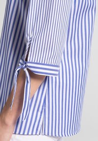 Blouse à manches courtes à rayures bleu clair et blanc, avec des poignets noués, des fentes latérales et un tissu lisse et léger.