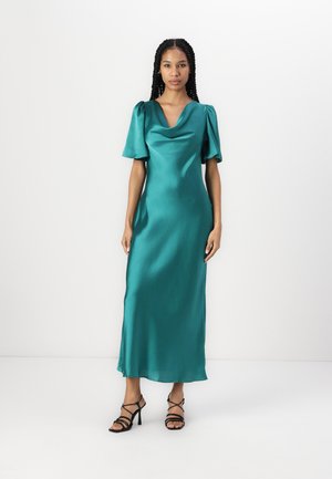Robe en satin teal avec un décolleté drapé, des manches courtes bouffantes et une silhouette fluide. Assortie à des sandales à talons hauts noires à lanières.