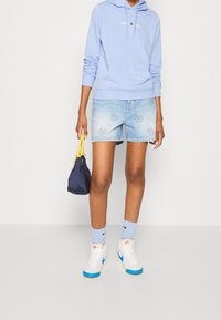 Sudadera con capucha azul claro con logotipo blanco, pantalones cortos de mezclilla con patrón de caritas sonrientes, zapatillas blancas con detalles azules y bolso pequeño oscuro.