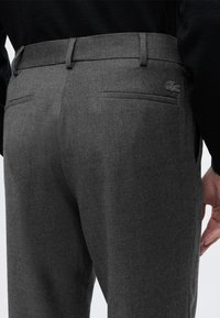 Pantalon gris foncé taillé avec passants de ceinture et poches passepoilées à l’arrière, avec un petit logo crocodile brodé sur la poche droite.