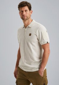 Polo shirt gris clair avec un col zippé, des manches courtes et une poche à rabat. Comprend un petit logo sur la poitrine et des accents sur les manches.