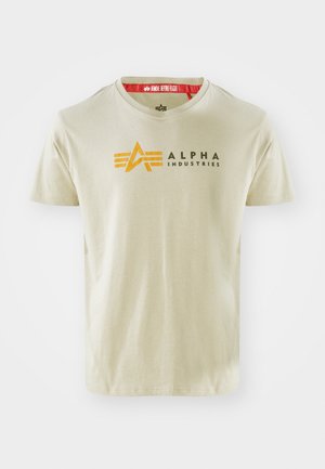 Vaaleanbeige puuvillainen t-paita lyhyillä hihoilla, jossa on musta ja oranssi "ALPHA INDUSTRIES" -logo ja suunnittelu yksityiskohtia rinnassa.