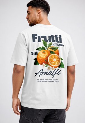 Mann trägt weißes T-Shirt mit Orangen-Grafik und Text „Frutti d'Italia Amalfi“ auf dem Rücken, schaut weg.