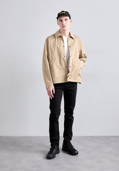 Veste beige à boutons, t-shirt blanc en dessous, pantalon noir et baskets noires. Casquette noire avec un logo jaune. Le mannequin se tient devant un fond neutre.