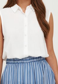 Vit ärmlös skjorta med knappar och krage, kombinerad med en blå och vit randig kjol med elastisk midja, som har vertikala veck.