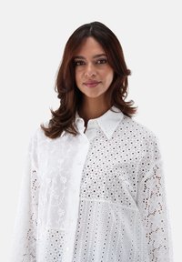Weißes Hemd mit Knopfleiste und verschiedenen Texturen, verziert mit floralem Stickerei und Lochmuster. Lange Ärmel, klassischer Kragen.