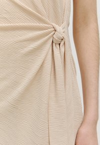 Beige strukturierter Stoff mit einem verdrehten Knoten-Detail, das Vorhänge bildet. Das Material weist vertikale Rippungen auf, die dem Design Tiefe verleihen.
