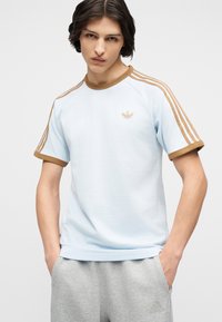 Νέος άνδρας με σκούρα μαλλιά που φοράει ένα ανοιχτό μπλε T-shirt Adidas με καφέ ρίγες και γκρι φόρμα Adidas, με τα χέρια στις τσέπες.