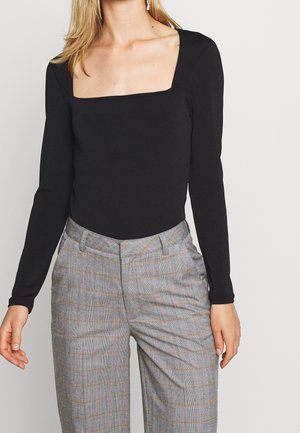 Femme portant un haut noir ajusté à manches longues avec un décolleté carré et un pantalon taille haute à carreaux gris avec des lignes orange.