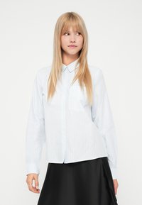 Svijetloplava košulja s dugim rukavima, okomitim prugama i ovratnikom. Ima džep na prsima i ležeran kroj. Uparena je s crnom suknjom.