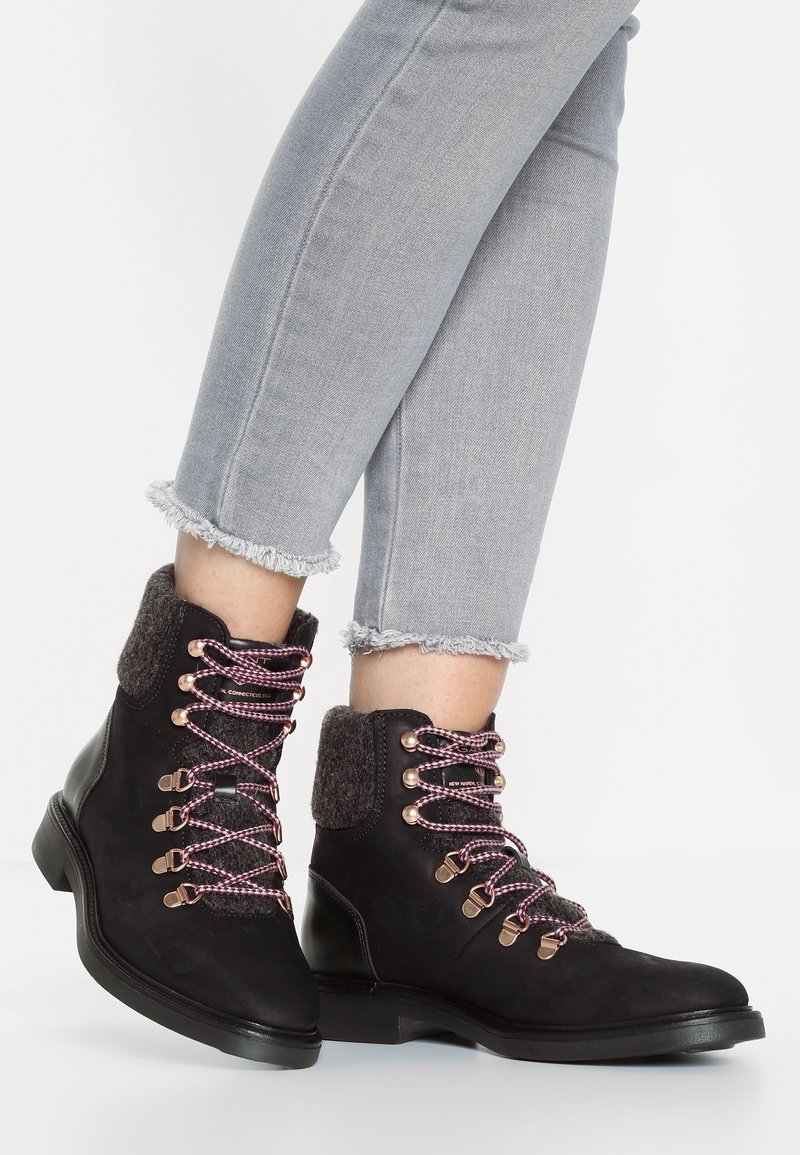 Schwarze knöchelhohe Schnürstiefel mit grauer Stoffauskleidung, pink- und grau gestreiften Schnürsenkeln und einer strukturierten Oberfläche. Flache Sohle mit minimalem Absatz.